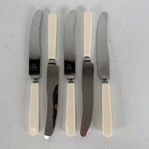 May include: A set of five stainless steel knives with smooth, cream-colored handles. Each blade features a stamped crown logo and the text "SOLINGEN ROSTFREI". The knives are arranged vertically, alternating the orientation of the blades and handles.