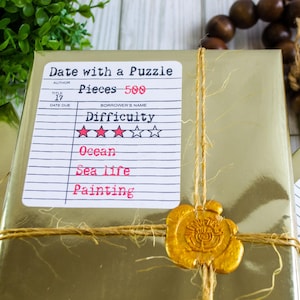 May include: A gold-wrapped gift box tied with twine and sealed with a gold wax stamp. A label reads "Date with a Puzzle," with details including 500 pieces, difficulty level, and themes like "Ocean" and "Sea life."