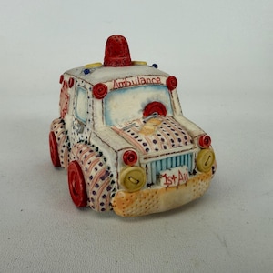 May include: A ceramic ambulance figurine featuring a white body with red and blue polka dot patterns. The vehicle has a red roof light, red wheels, and yellow headlights. The word Ambulance is written above the windshield, and 1st Aid is on the front bumper.