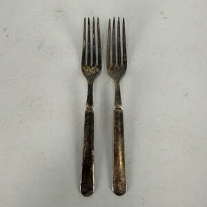 May include: Two silver-toned metal forks with four tines each. The handles are straight and rectangular with a small, embossed shell-like motif at the base. The metal surface shows signs of oxidation and tarnishing, giving it a dark, aged appearance.