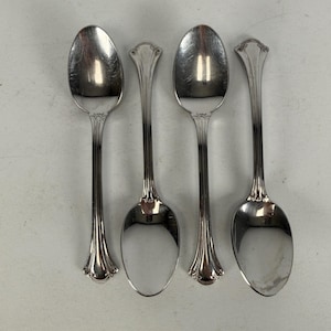 May include: A set of four silver-toned metal spoons arranged in a row. Each spoon features a polished oval bowl and a slender handle with a vertical grooved pattern and a decorative, rounded scroll-like detail at the top of the handle stem.