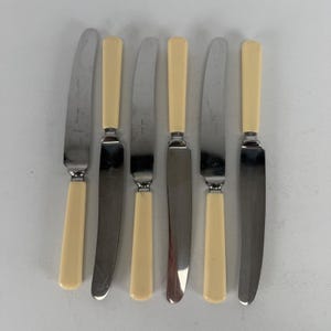 May include: A set of six table knives with polished silver-tone metal blades and smooth, cream-colored handles. The knives are arranged in a vertical row, alternating the orientation of the blades and handles to create a uniform appearance.
