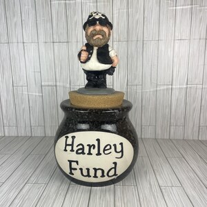 May include: A ceramic jar with a cork lid topped by a figurine of a bearded person wearing a black vest, black pants, and a black cap with a skull emblem. The jar is dark gray with a white oval label on the front featuring the black text Harley Fund.