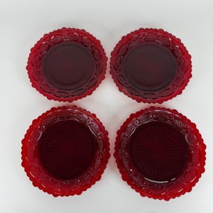 May include: A set of four circular red glass plates featuring a scalloped edge and a decorative embossed pattern along the rim. Each plate has a central sunburst design with radiating lines, crafted from translucent deep red pressed glass.