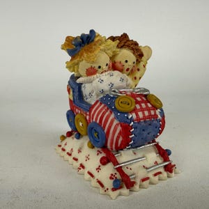 May include: A decorative figurine featuring two doll-like figures with blonde and brown hair riding in a train car. The train is decorated with red and white stripes, blue patches, and yellow buttons. It sits on a white base with red stitching details.