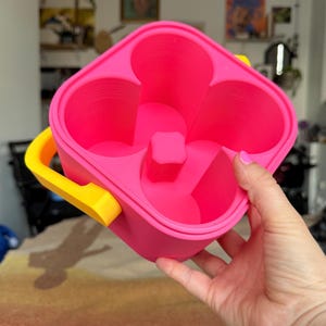 Puede incluir: Un portavasos rosa brillante con un asa amarilla. El portavasos tiene cuatro portavasos y un divisor central. El asa est&aacute; unida al lateral. El portavasos lo sostiene una persona.