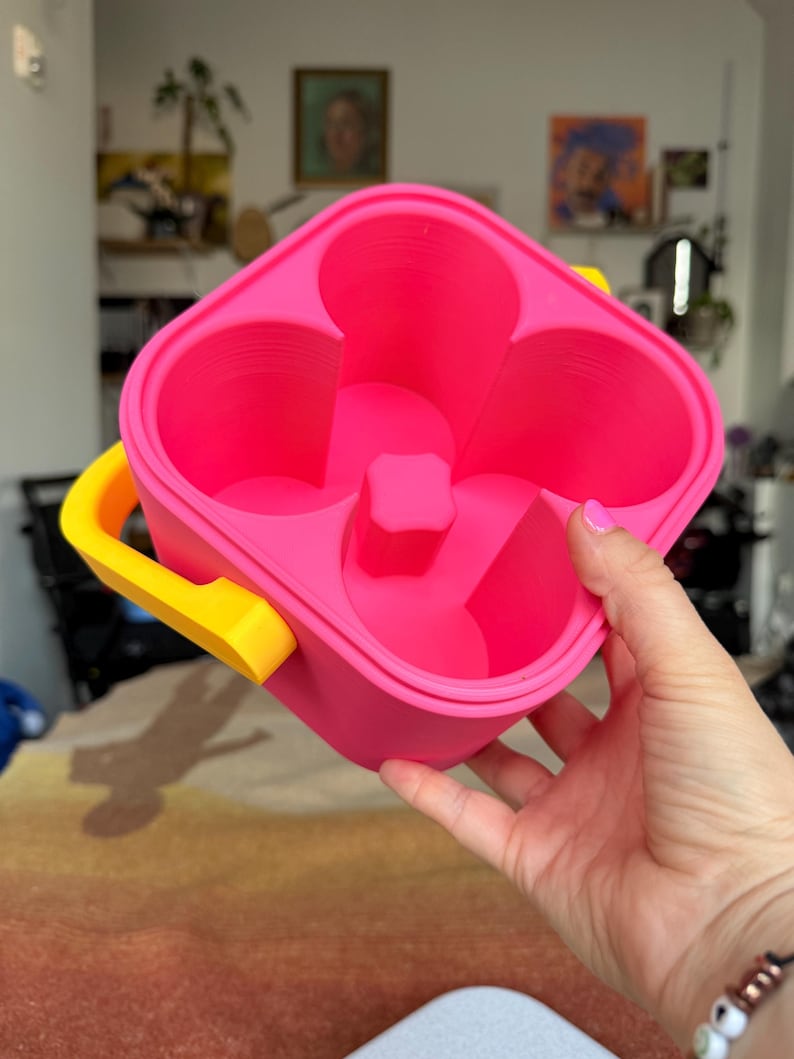 Puede incluir: Un portavasos rosa brillante con un asa amarilla. El portavasos tiene cuatro portavasos y un divisor central. El asa est&aacute; unida al lateral. El portavasos lo sostiene una persona.