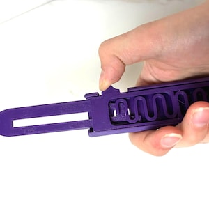 May include: A purple key-shaped tool with a slotted blade and a wavy interior design. The tool is held in a hand against a white background. The tool appears to be made of plastic.