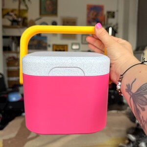 Puede incluir: Una nevera rosa brillante con una tapa gris moteada y un asa amarilla. La nevera es sostenida por una persona. El fondo incluye obras de arte y una alfombra estampada.