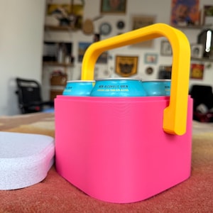 Puede incluir: Un portabebidas rosa brillante con un asa amarilla contiene varias latas azules. Las latas muestran el texto "NON-ALCOHOLIC BREW" y "CONTAINS LESS THAN 0.5% ALCOHOL". Un posavasos blanco est&aacute; situado a la izquierda del portabebidas.