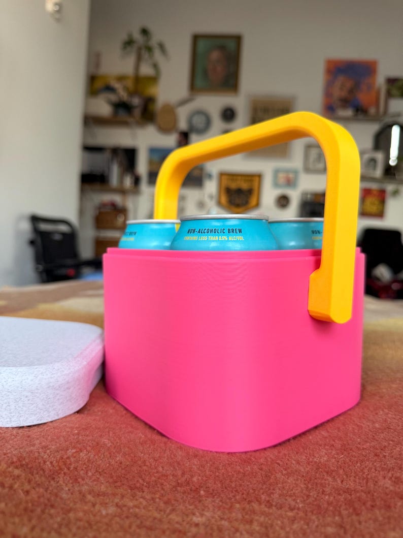 Puede incluir: Un portabebidas rosa brillante con un asa amarilla contiene varias latas azules. Las latas muestran el texto "NON-ALCOHOLIC BREW" y "CONTAINS LESS THAN 0.5% ALCOHOL". Un posavasos blanco est&aacute; situado a la izquierda del portabebidas.