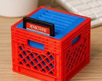 May include: A red and blue SD card holder shaped like a miniature crate. The holder has a red exterior and a blue interior, with a SanDisk SD card inserted. The design is reminiscent of a small storage container.