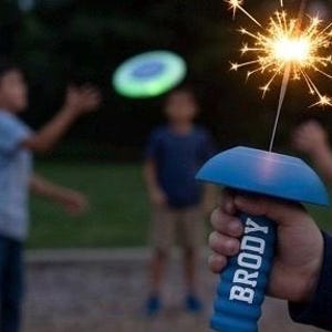 Puede incluir: Un soporte azul para bengalas con el nombre "BRODY" en letras blancas se sostiene en la mano de una persona. Una bengala encendida emite chispas brillantes. Dos frisbees brillantes están en el aire, con niños en el fondo.