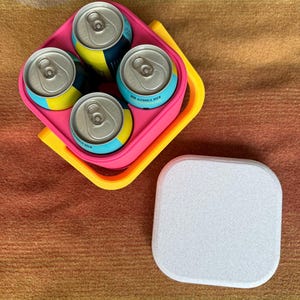 Puede incluir: Un portabebidas rosa y amarillo que contiene cuatro latas de cerveza sin alcohol. Las latas tienen etiquetas azules y amarillas. Una posavasos blanco y rectangular est&aacute; al lado del portabebidas sobre una superficie marr&oacute;n.