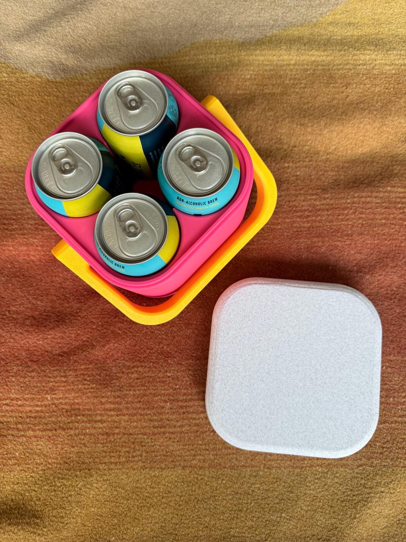 Puede incluir: Un portabebidas rosa y amarillo que contiene cuatro latas de cerveza sin alcohol. Las latas tienen etiquetas azules y amarillas. Una posavasos blanco y rectangular est&aacute; al lado del portabebidas sobre una superficie marr&oacute;n.