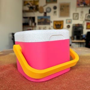 Puede incluir: Una nevera rosa brillante con una tapa blanca moteada y un asa amarilla. La nevera tiene forma cuadrada con esquinas redondeadas. El asa es arqueada y est&aacute; hecha de un material amarillo s&oacute;lido.