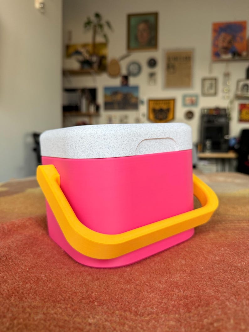 Puede incluir: Una nevera rosa brillante con una tapa blanca moteada y un asa amarilla. La nevera tiene forma cuadrada con esquinas redondeadas. El asa es arqueada y est&aacute; hecha de un material amarillo s&oacute;lido.