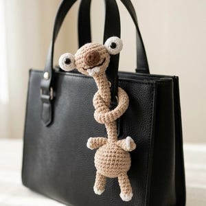 Pode incluir: Uma mala de mão de couro preta com um pingente de crochê. O pingente é uma criatura marrom clara com olhos brancos grandes e um rosto sorridente. O pingente está preso à alça da mala.