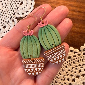 May include: A pair of handmade cactus earrings. The earrings feature green cactus shapes with pink flower accents, set in brown pots with white geometric designs. The earrings have gold-colored ear wires.