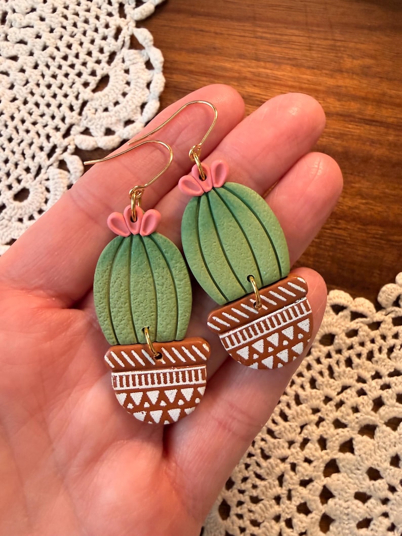 May include: A pair of handmade cactus earrings. The earrings feature green cactus shapes with pink flower accents, set in brown pots with white geometric designs. The earrings have gold-colored ear wires.