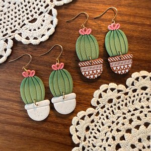 May include: Handmade cactus earrings with gold-colored hooks. The earrings feature green cactus shapes with pink flower accents, set in white or brown patterned pots. The earrings are displayed on a wooden surface with white lace doilies.