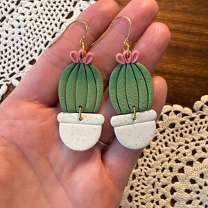 May include: A pair of handmade cactus earrings. Each earring features a green cactus with pink flower accents, set in a white speckled pot. The earrings have gold-colored hooks for pierced ears. The earrings are a unique, nature-inspired accessory.