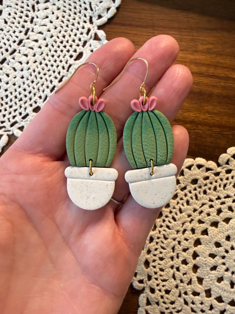 May include: A pair of handmade cactus earrings. Each earring features a green cactus with pink flower accents, set in a white speckled pot. The earrings have gold-colored hooks for pierced ears. The earrings are a unique, nature-inspired accessory.
