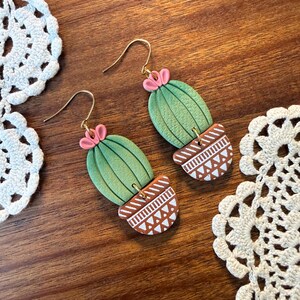 May include: A pair of handmade cactus earrings. The earrings feature a green cactus design with pink flower accents, set in a brown pot with white geometric patterns. The earrings have gold-colored hooks.