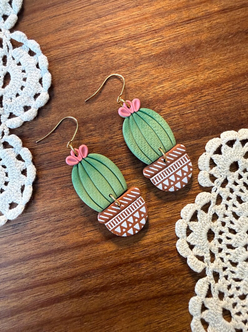 May include: A pair of handmade cactus earrings. The earrings feature a green cactus design with pink flower accents, set in a brown pot with white geometric patterns. The earrings have gold-colored hooks.