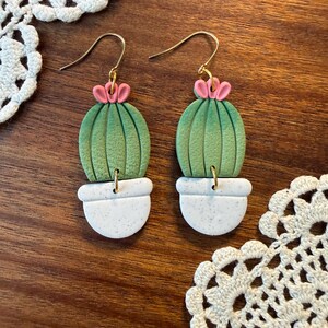 May include: A pair of handmade earrings featuring a cactus design. Each earring has a green cactus with pink flower accents, set in a white speckled pot. The earrings hang from gold-colored ear wires. The earrings are displayed on a wooden surface.