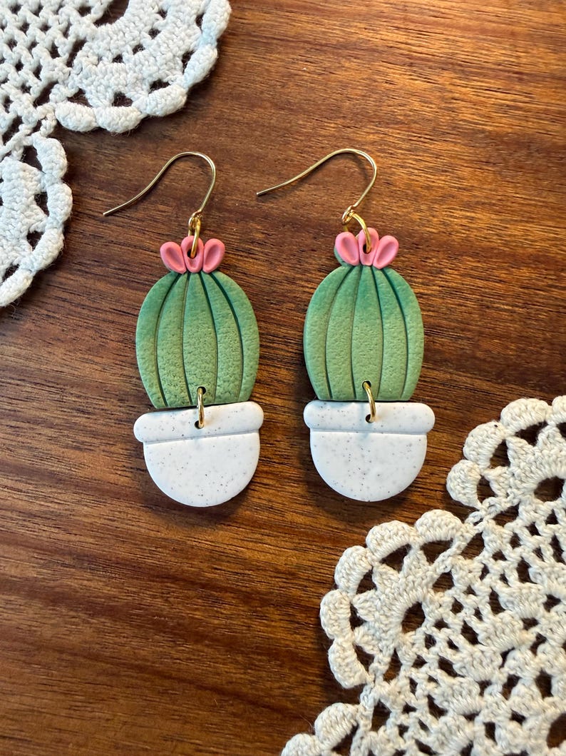 May include: A pair of handmade earrings featuring a cactus design. Each earring has a green cactus with pink flower accents, set in a white speckled pot. The earrings hang from gold-colored ear wires. The earrings are displayed on a wooden surface.