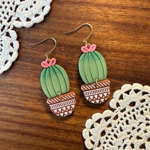 May include: A pair of handmade earrings featuring a cactus design. The earrings have a green cactus body with pink flower accents and are set in a brown pot with white geometric patterns. The earrings hang from gold-colored hooks.