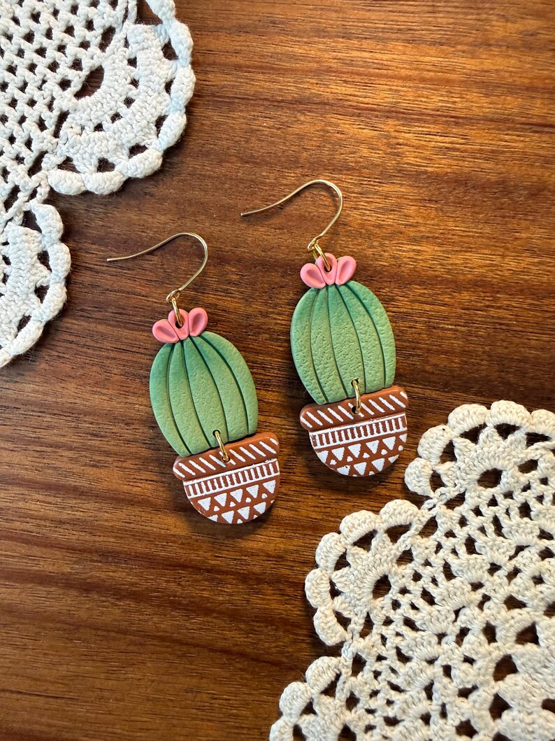 May include: A pair of handmade earrings featuring a cactus design. The earrings have a green cactus body with pink flower accents and are set in a brown pot with white geometric patterns. The earrings hang from gold-colored hooks.