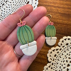 May include: Handmade cactus earrings. Each earring features a green cactus with pink flower accents, set in a white speckled pot. The earrings have gold-colored hooks. The background includes a wooden surface and a white lace doily.