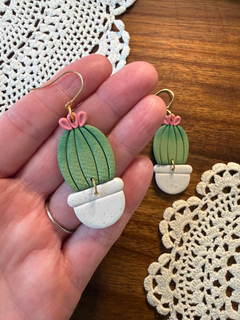 May include: Handmade cactus earrings. Each earring features a green cactus with pink flower accents, set in a white speckled pot. The earrings have gold-colored hooks. The background includes a wooden surface and a white lace doily.