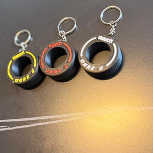 Könnte beinhalten: Drei schwarze, reifenförmige Schlüsselanhänger mit silbernen Schlüsselringen. Jeder Schlüsselanhänger hat ein andersfarbiges Band: gelb, rot und weiß. Der Text "P ZERO" ist auf jedem Reifen sowie das Pirelli-Logo zu sehen.