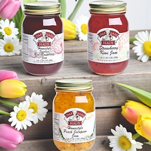 May include: Three glass jars of Das Haus jam with gold lids, each labeled with a different flavor: Seedless Red Raspberry, Strawberry Kiwi, and Peach Jalapeno. The jars are arranged on a wooden surface, surrounded by flowers, and the text "Choose Any 3 Flavors" is at the top.