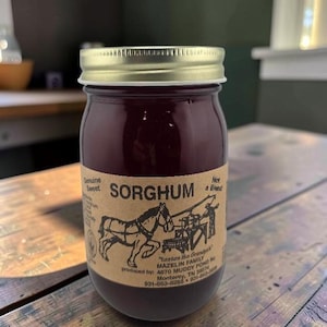 May include: A glass jar filled with dark amber sorghum syrup, topped with a gold metal lid. The tan paper label features a black illustration of a horse-drawn mill and text reading Genuine Sweet SORGHUM Not a Blend, Mazelin Family, and address.