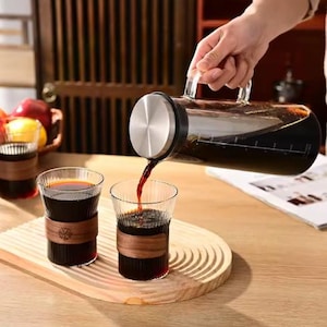 May include: A clear glass carafe with a stainless steel lid is pouring a dark liquid into two ribbed glasses. The glasses have wooden sleeves. The carafe and glasses are on a wooden tray.