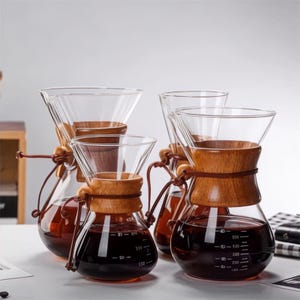 May include: Four glass coffee makers with wooden collars and leather ties. The coffee makers have a clear, conical top and a rounded bottom, filled with dark liquid. The wooden collars are secured with leather ties. The coffee makers are on a white surface.