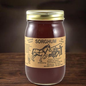 May include: A glass jar filled with dark amber sorghum syrup, topped with a gold metal lid. The tan label features a black illustration of a horse-drawn press, the text SORGHUM, and production details including the Mazelin Family address.