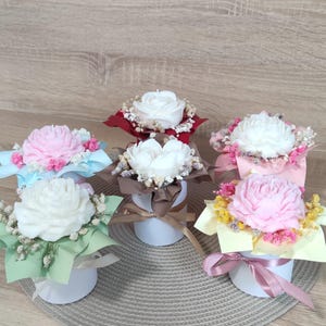 Op de afbeelding: Zes kleine, witte cilindrische containers die elk een bloemvormige kaars bevatten. De kaarsen zijn wit en roze, omringd door decoratief papier, gedroogde bloemen en linten. De opstelling staat op een lichtgekleurd houten oppervlak.