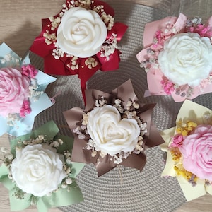 Könnte beinhalten: Sechs Blumenarrangements, jedes mit einer geformten Rose im Mittelpunkt. Die Rosen sind weiß und rosa, umgeben von farbigem Papier und Trockenblumen. Die Arrangements werden auf einer hellen Oberfläche präsentiert. Jedes Arrangement ist einzigartig.
