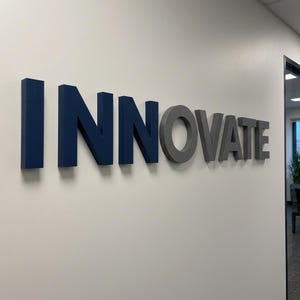 May include: A three-dimensional wall sign featuring the word INNOVATE in block lettering. The letters I, N, N, and O are dark blue, while the letters V, A, T, and E are a neutral gray. The sign is mounted on a smooth, off-white interior wall.