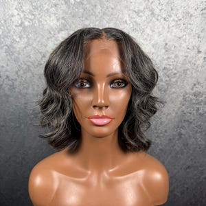 May include: A gray and black wavy wig styled on a mannequin head. The wig features a natural-looking hairline and a short, layered cut. The mannequin has brown skin, brown eyes, and pink lipstick. The background is a mottled gray.