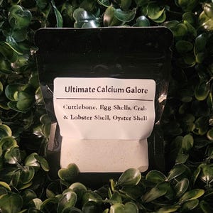 May include: A black resealable pouch containing a calcium supplement powder. The white label reads Ultimate Calcium Galore and lists the ingredients as Cuttlebone, Egg Shells, Crab & Lobster Shell, and Oyster Shell in black text.
