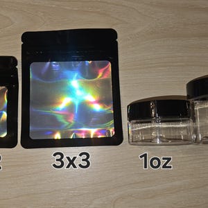 May include: Two black pouches with holographic windows labeled 2x2 and 3x3, alongside two clear plastic jars with black lids labeled 1oz and 2oz. The pouches feature a zip-top closure, and the jars are cylindrical with a transparent body.