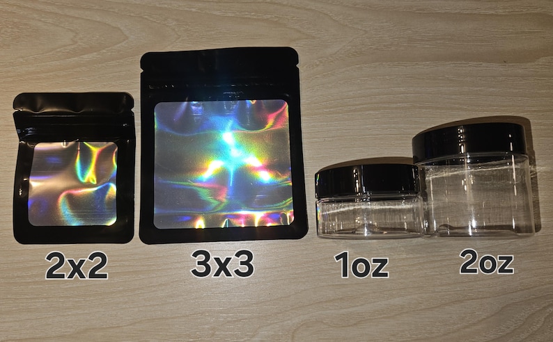 May include: Two black pouches with holographic windows labeled 2x2 and 3x3, alongside two clear plastic jars with black lids labeled 1oz and 2oz. The pouches feature a zip-top closure, and the jars are cylindrical with a transparent body.