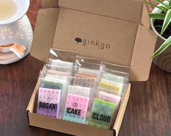 May include: A brown cardboard box labeled ginkgo containing multiple rectangular wax melts in clear plastic sleeves. The front row features three wax melts with the text Sugar, Cake, and Cloud printed in black on pink, blue, and green backgrounds.
