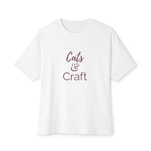 May include: White t-shirt with the text "Cats & Craft" in a burgundy color. The shirt has short sleeves and a crew neck. The text is in a handwritten style font.
